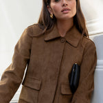Woman wearing a brown suede jacket and blue jeans sitting on a white surface.