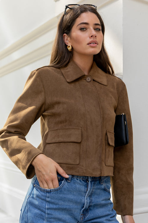 Woman wearing a brown jacket and blue jeans against a white wall.