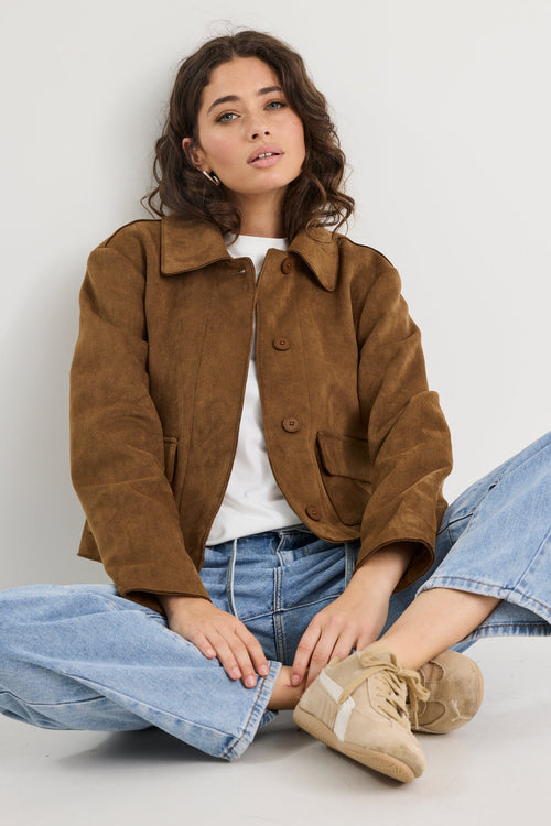 Woman wearing a brown jacket, white shirt, blue jeans, and beige boots sitting on the floor against a white background.