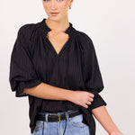A woman modeling a black pinstriped blouse with a gathered ruffled neckline and voluminous elbow-length sleeves, paired with light blue jeans and a black belt, against a plain light background.