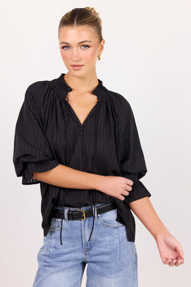 A woman modeling a black pinstriped blouse with a gathered ruffled neckline and voluminous elbow-length sleeves, paired with light blue jeans and a black belt, against a plain light background.