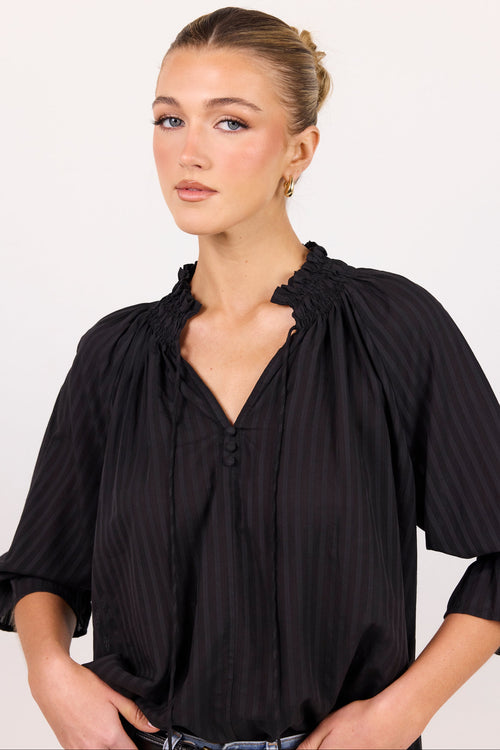 A woman wearing a black pleated blouse with a gathered ruffled collar and three-quarter sleeves, photographed against a plain light background.