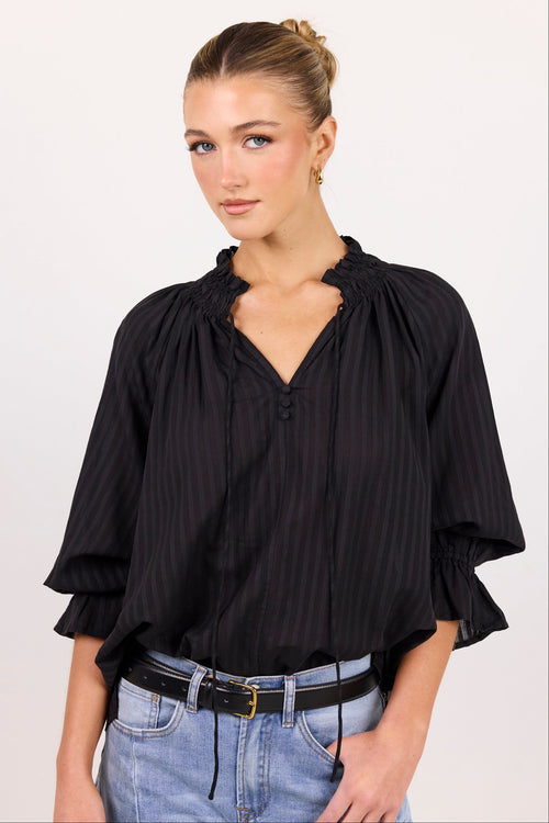 A woman modeling a black and navy vertically striped blouse with ruffled trim and elbow-length sleeves, paired with light blue jeans and a black belt, against a plain light background.
