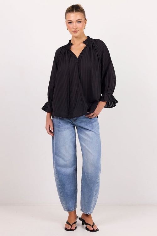 A woman modeling a black pleated long-sleeve blouse with ruffled cuffs, paired with light-wash blue jeans and black sandals, standing against a plain white studio background.