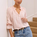 A woman wearing a light pink ruffled blouse and blue jeans stands in a bright indoor setting with white walls and wooden stairs.