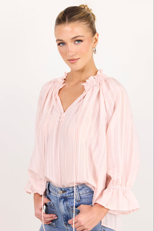 A woman modeling a light pink long-sleeve blouse with ruffled trim and drawstring ties, paired with blue jeans, against a plain white studio background.