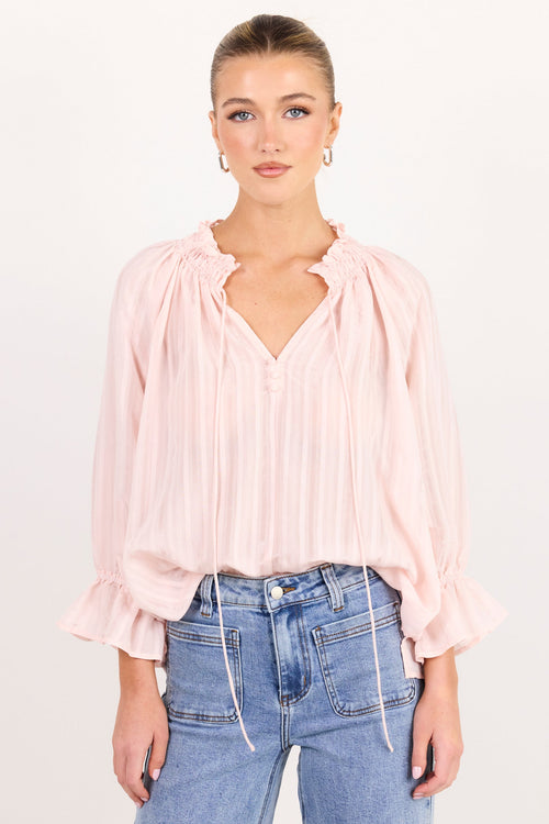 A woman modeling a light pink pleated blouse with a split neckline and ruffled cuffs, paired with light blue jeans, against a plain white studio background.