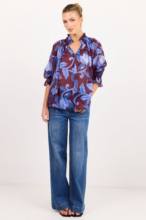 A woman modeling a long-sleeve blouse with a blue and burgundy floral pattern, paired with wide-leg blue jeans and black sandals, standing against a plain white studio background.