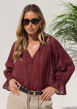 A woman wearing a burgundy long-sleeve blouse with a V-neck and gathered sleeves, paired with beige skirt and black sunglasses, standing against a light wall with a plant in the background.