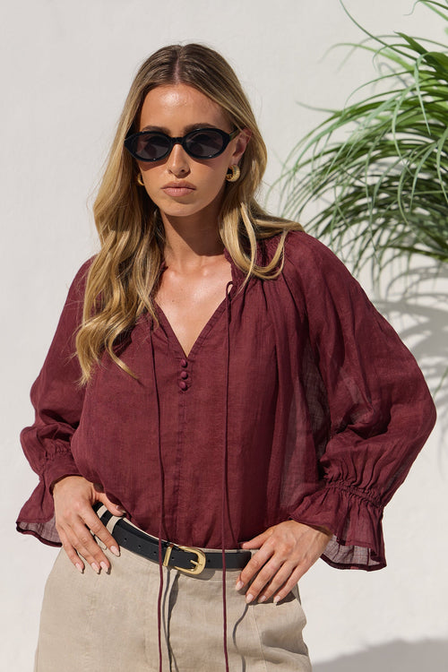 A woman wearing a burgundy long-sleeve blouse with a V-neck and gathered sleeves, paired with beige skirt and black sunglasses, standing against a light wall with a plant in the background.