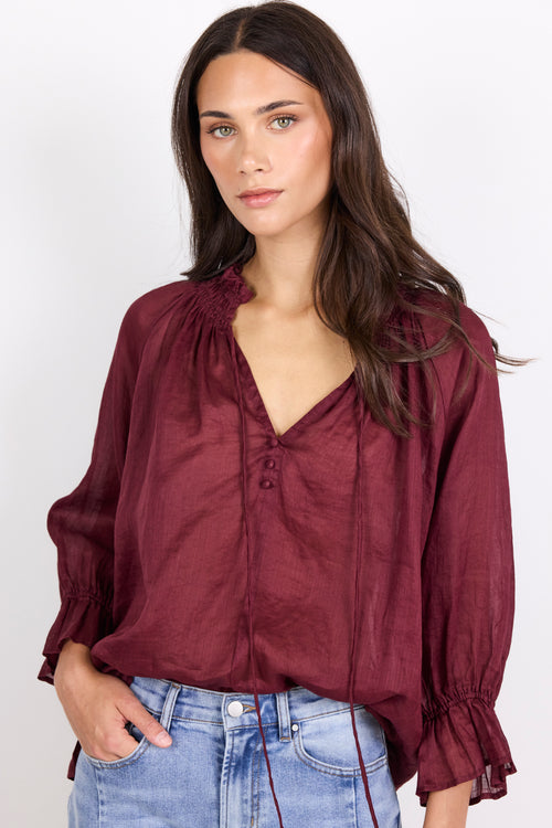 A woman wearing a burgundy long-sleeve blouse with a gathered V-neckline and ruffled cuffs, paired with light blue jeans, against a plain white background.