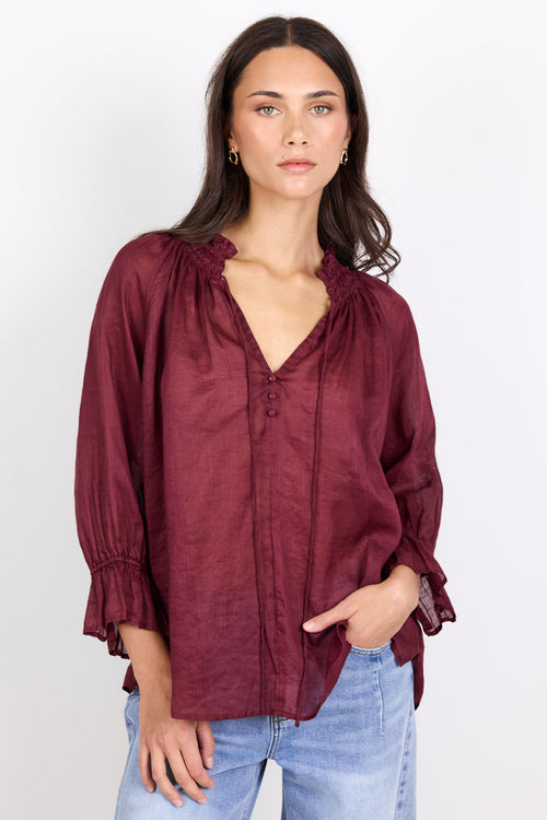A woman wearing a burgundy long-sleeve blouse with a gathered ruffled neckline and button front, paired with light blue jeans, standing against a plain white studio background.