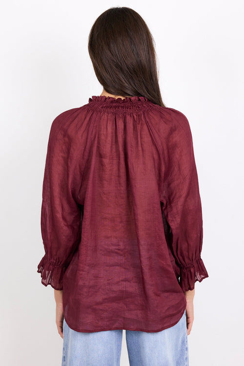 A woman viewed from the back wearing a long-sleeve burgundy blouse with a smocked neck and ruffled cuffs, paired with light blue jeans, against a plain white background.
