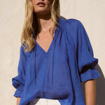 Woman wearing a blue blouse and white pants against a beige wall