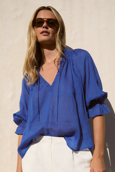 Woman wearing a blue blouse and white pants against a beige wall