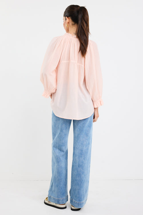 woman wearing a soft pink flowy cotton blouse with tie neckline and frill details, paired with blue jeans.