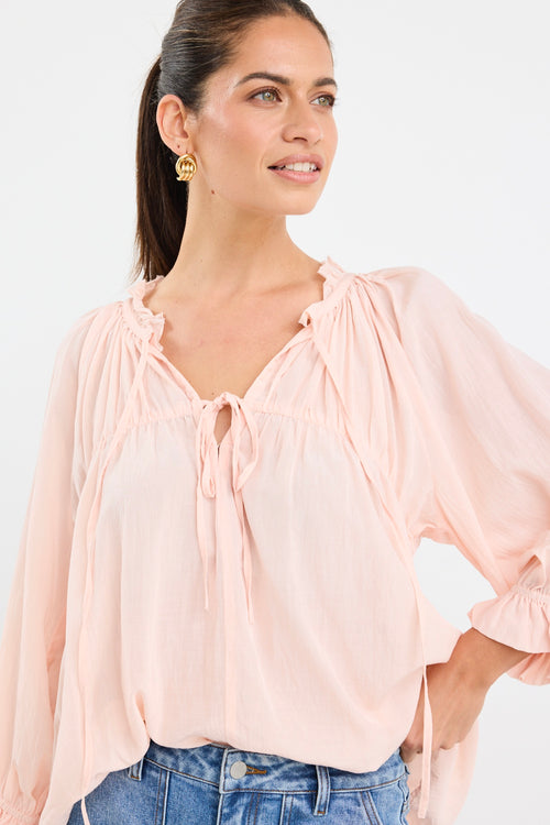 woman wearing a soft pink flowy cotton blouse with tie neckline and frill details, paired with blue jeans.