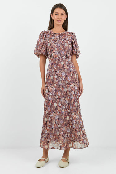 A woman wearing a floral midi dress in shades of lilac, blush, and soft pink, with puffed short sleeves and a relaxed fit.