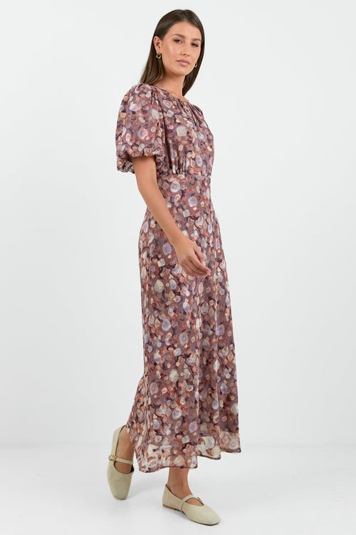 A woman wearing a floral midi dress in shades of lilac, blush, and soft pink, with puffed short sleeves and a relaxed fit.