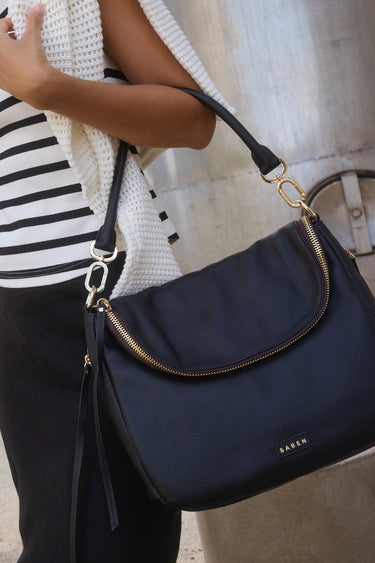 Angled front view of a black pebbled leather shoulder bag featuring a soft fold-over flap with gold zip detailing. The structured shape is complemented by gold hardware and a detachable top handle. The brand name 'SABEN' is embossed in gold at the base front of the bag.