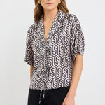 Woman wearing a short sleeve satin shirt with bold black geometric print, button front, and tie waist detail, styled with black wide-leg jeans and white sandals.