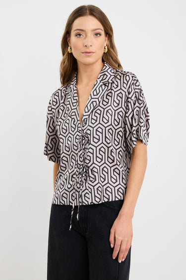 Woman wearing a short sleeve satin shirt with bold black geometric print, button front, and tie waist detail, styled with black wide-leg jeans and white sandals.