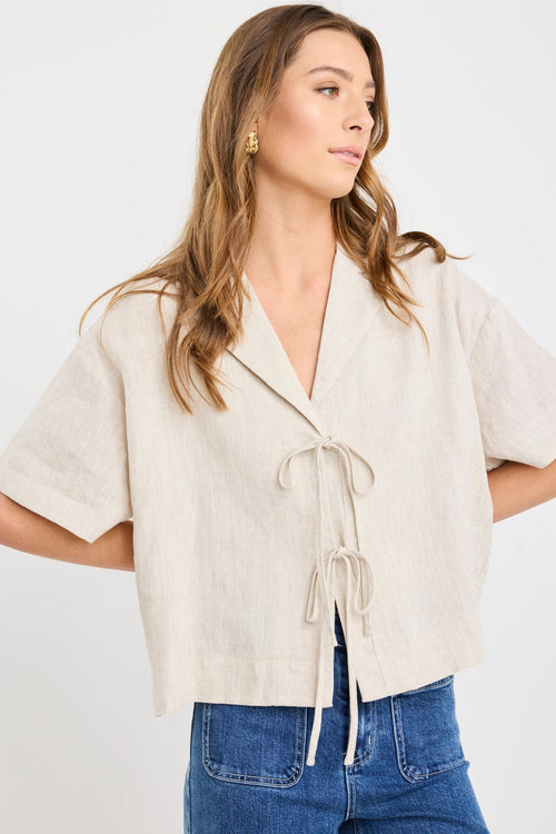Woman wearing a natural beige short sleeve linen shirt with tie-front fastening, styled with mid-blue jeans.