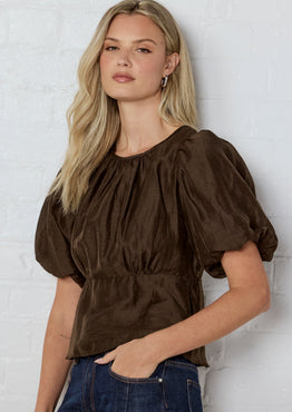 Woman wearing a brown blouse with puffed sleeves against a white brick wall.