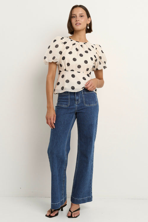 Woman wearing a cream short sleeve top with black polka dots, puff sleeves, and blue jeans.