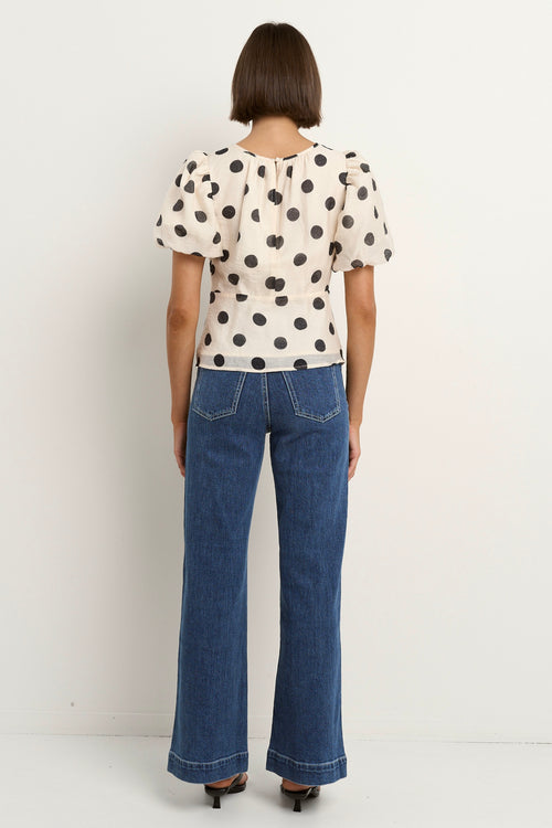 Woman wearing a cream short sleeve top with black polka dots, puff sleeves, and blue jeans.