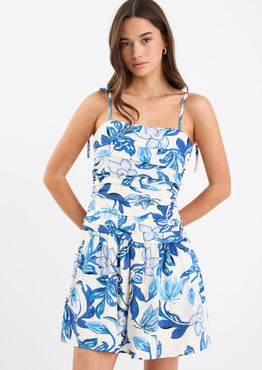 Woman wearing a blue floral mini dress with a pleated bodice, strappy sleeves, and a flattering fit, paired with white sandals.