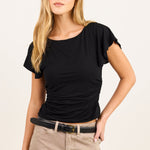 A blonde woman modeling a black short-sleeve top with ruching, paired with beige pants and a black belt, against a plain off-white studio background.