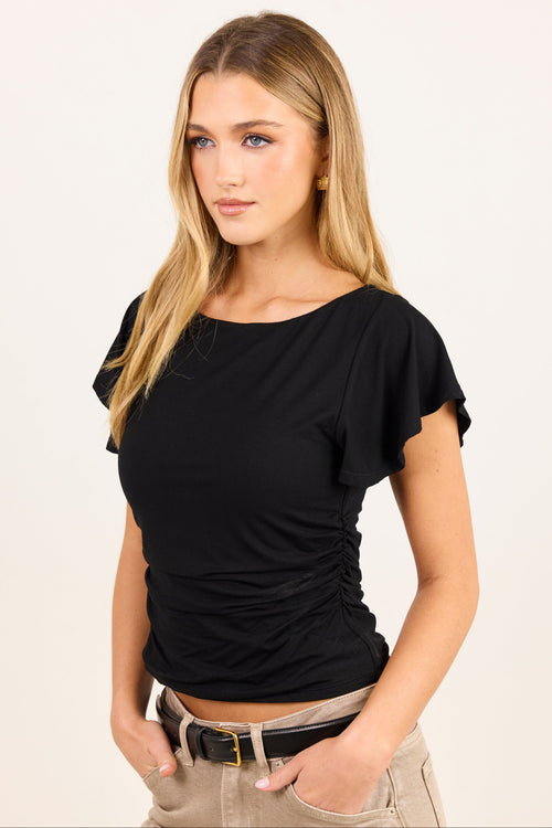 A woman modeling a black short-sleeve top with flutter sleeves and a gathered ruched waist, paired with beige pants, against a plain light background.