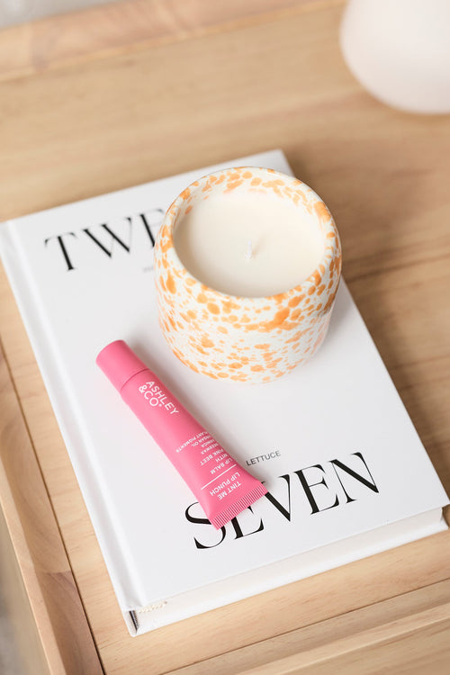 A styled product shot featuring a pink lip balm tube and a patterned candle on a white book, set on a light wood tray next to a white table lamp.