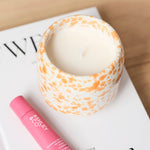 A styled product shot featuring a pink lip balm tube and a patterned candle on a white book, set on a light wood tray next to a white table lamp.