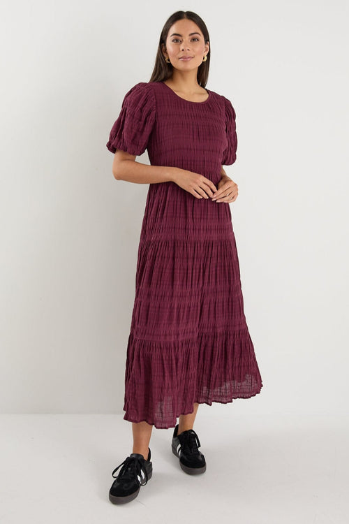 Woman wearing a plum puff-sleeve maxi dress with a textured finish and tiered skirt, paired with black sneakers.