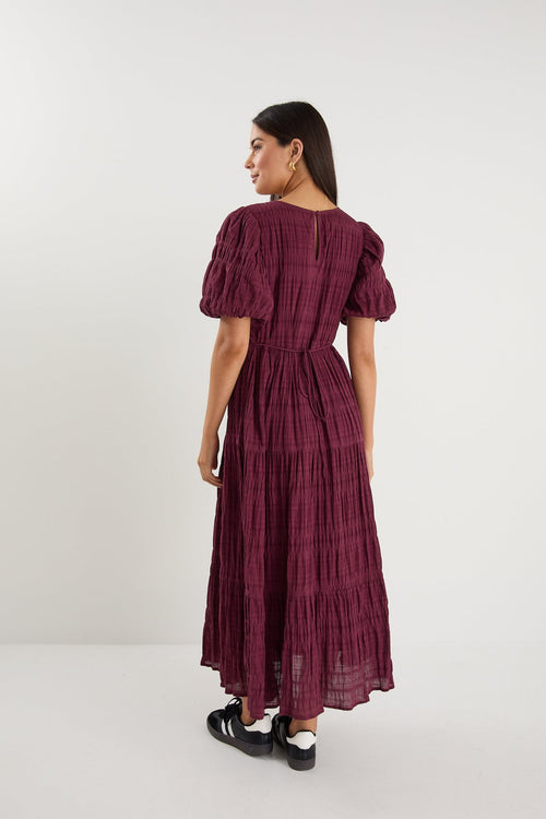 Woman wearing a plum puff-sleeve maxi dress with a textured finish and tiered skirt, paired with black sneakers.