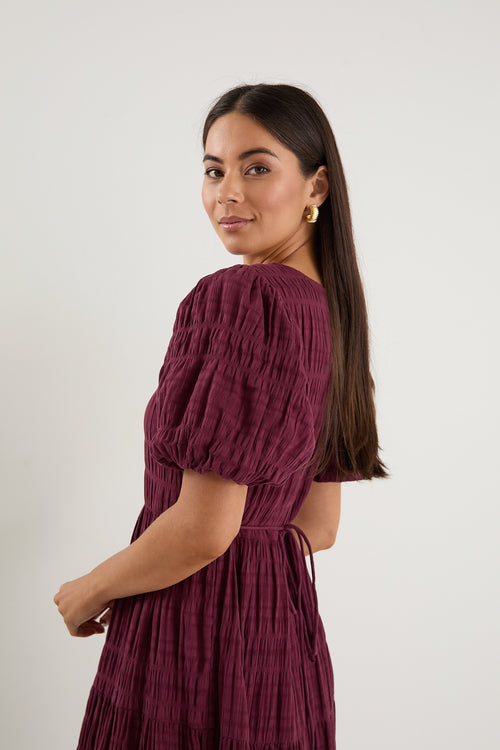 Woman wearing a plum puff-sleeve maxi dress with a textured finish and tiered skirt, paired with black sneakers.