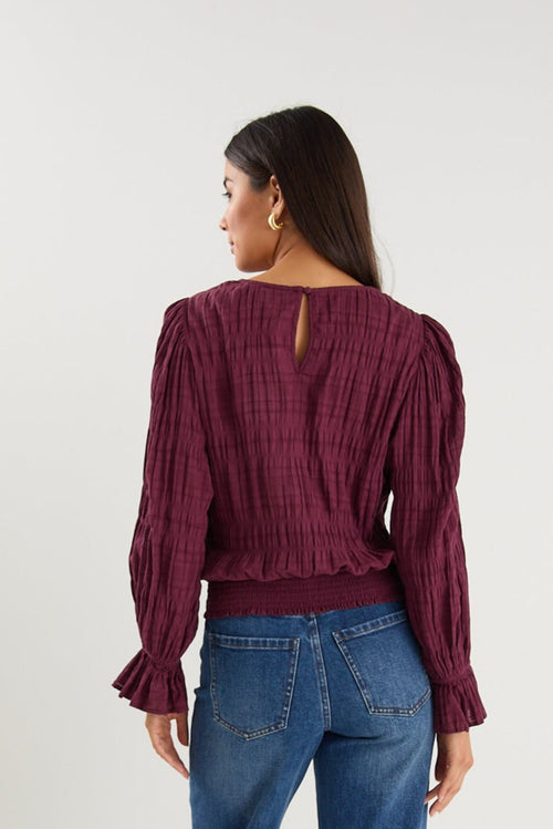 Woman wearing a long-sleeve textured blouse in deep plum, styled with blue high-waisted jeans and gold hoop earrings.