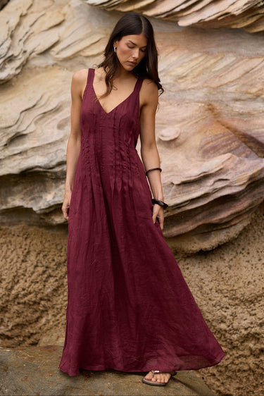 A woman wearing a sleeveless, V-neck maxi dress in deep burgundy stands on a rock in front of a large layered sandstone cliff.