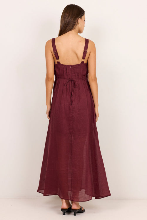 A woman viewed from the back wearing a sleeveless burgundy maxi dress with thin shoulder straps, a gathered waist, and a tie detail, standing against a plain light background.