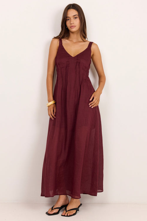 A woman modeling a sleeveless, V-neck maxi dress in a deep burgundy color, standing against a plain white studio background.