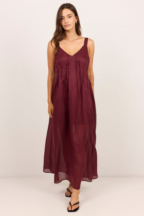 A woman modeling a sleeveless, V-neck maxi dress in a deep burgundy color, standing against a plain off-white studio background.