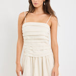 Woman wearing a plain ecru mini dress with thin spaghetti straps, a pleated bodice, and softly flared skirt.