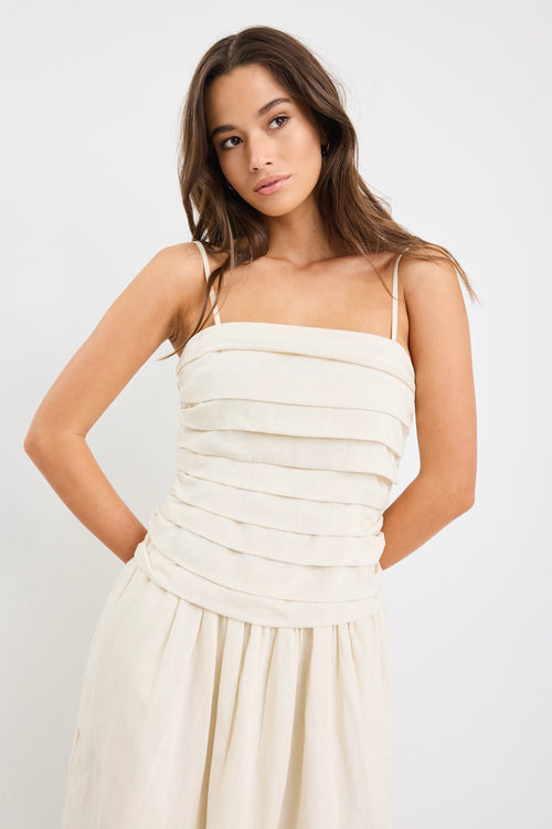 Woman wearing a plain ecru mini dress with thin spaghetti straps, a pleated bodice, and softly flared skirt.