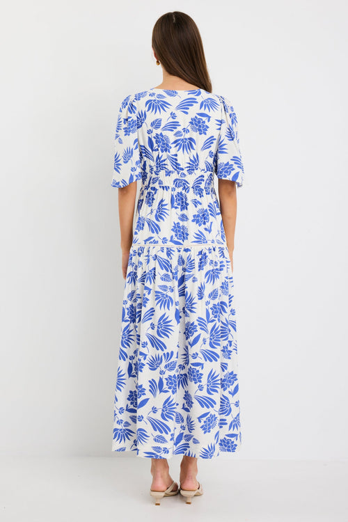Woman wearing a white maxi dress with bold blue floral print, flutter sleeves, v-neckline, and tiered skirt, styled with neutral sandals.
