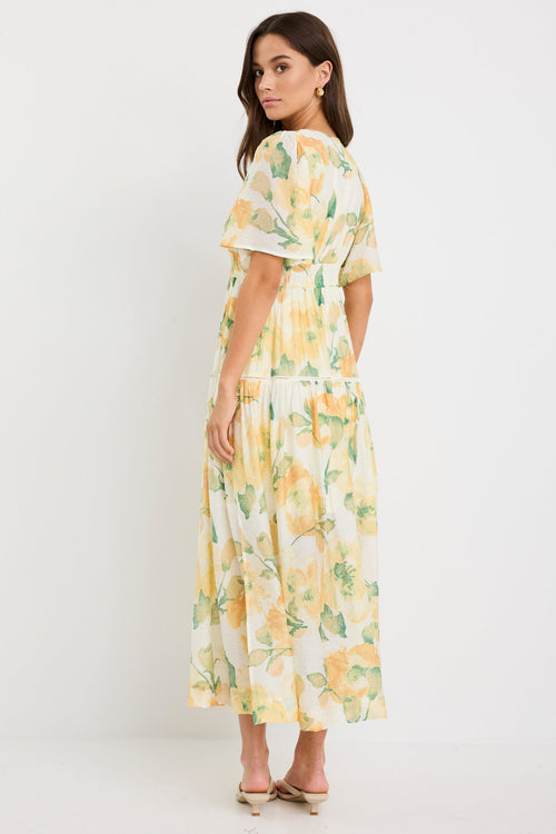 Woman wearing a honey-yellow floral maxi dress with flutter sleeves, v-neckline, and tiered flowing skirt, styled with neutral sandals.