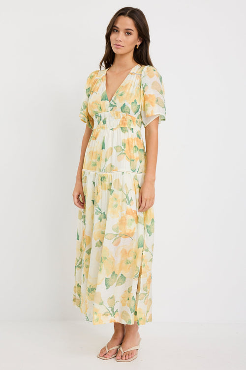 Woman wearing a honey-yellow floral maxi dress with flutter sleeves, v-neckline, and tiered flowing skirt, styled with neutral sandals.