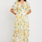Woman wearing a honey-yellow floral maxi dress with flutter sleeves, v-neckline, and tiered flowing skirt, styled with neutral sandals.