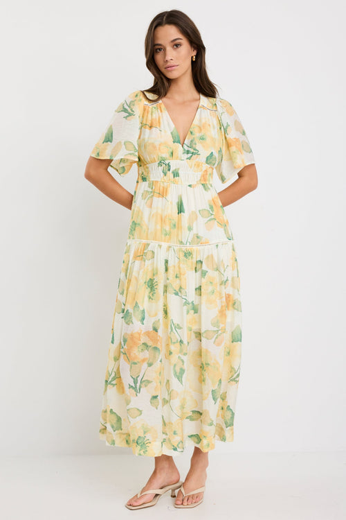 Woman wearing a honey-yellow floral maxi dress with flutter sleeves, v-neckline, and tiered flowing skirt, styled with neutral sandals.